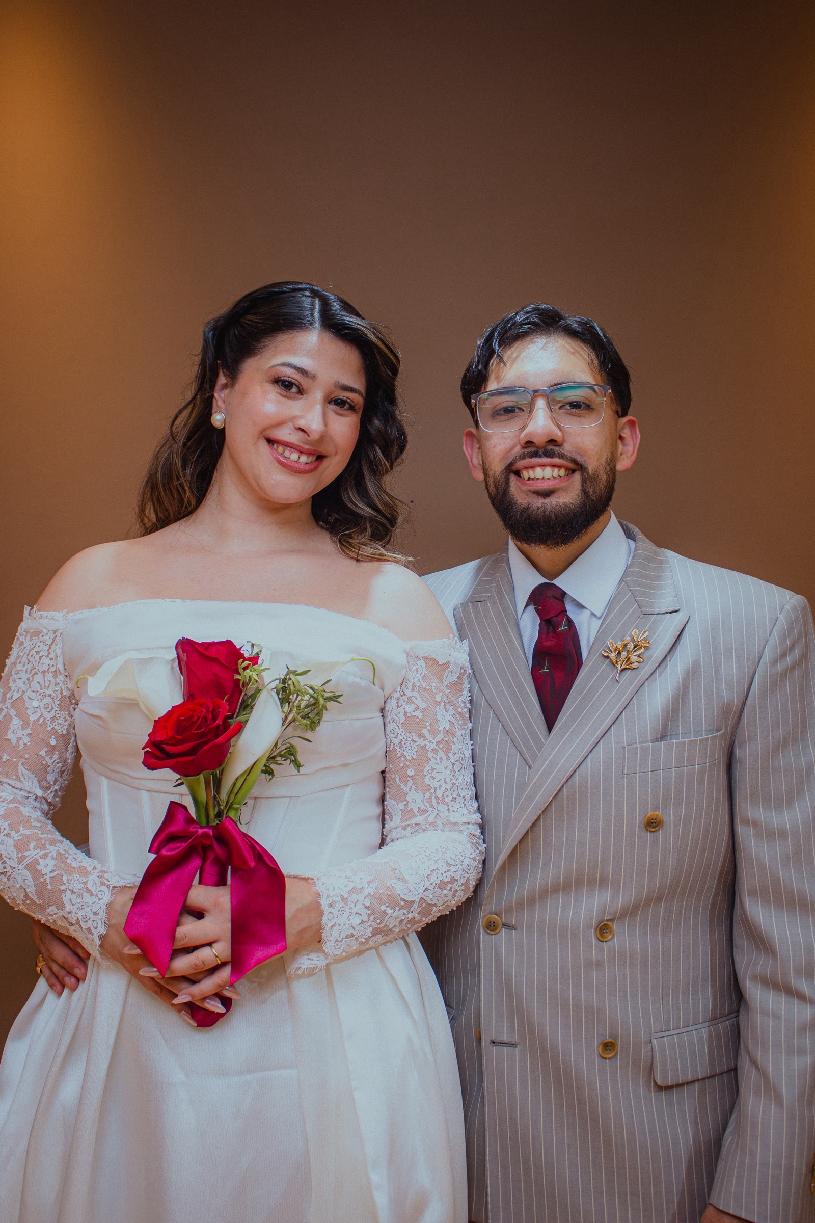 Luanne y Ramiro sonriendo el día de su boda, ella con un ramo de rosas rojas y calas